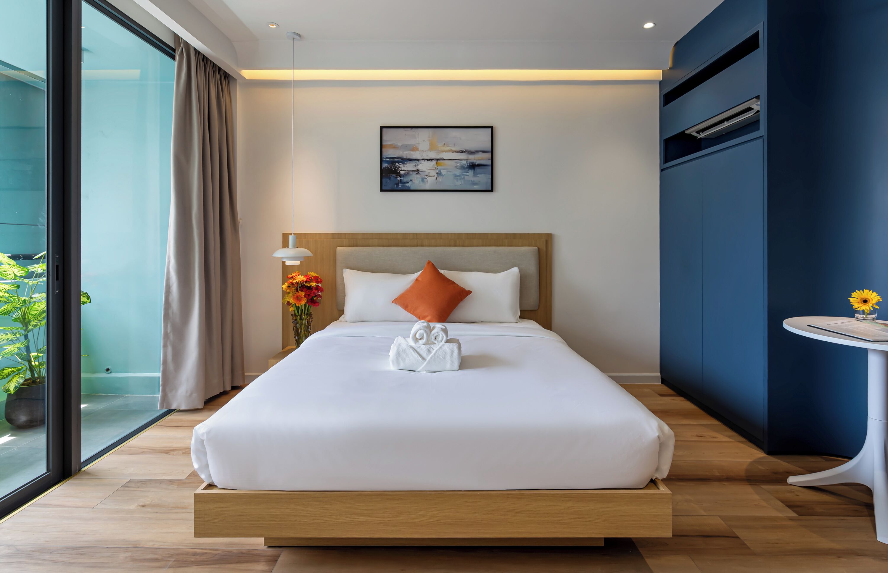 Une chambre d’hôtel moderne avec un grand lit, une table de chevet ornée d’un vase de fleurs, un tableau au mur et une vue dégagée sur l’extérieur grâce à une grande fenêtre.