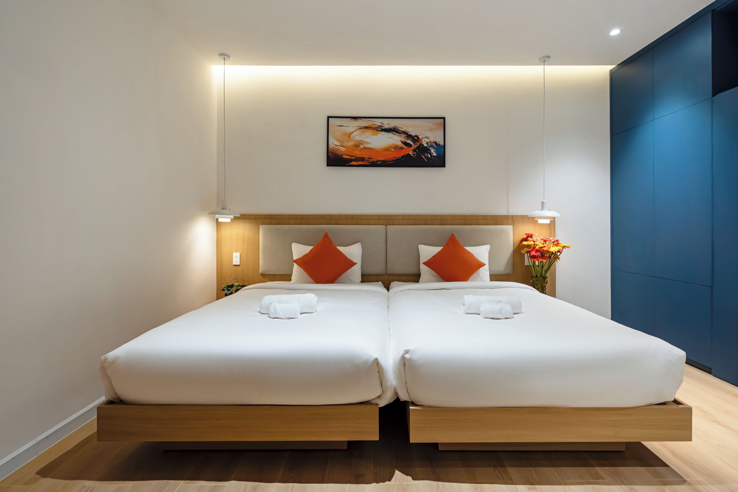 A double bed with white bedding and orange pillows, a painting above, and a vase of flowers on the nightstand.