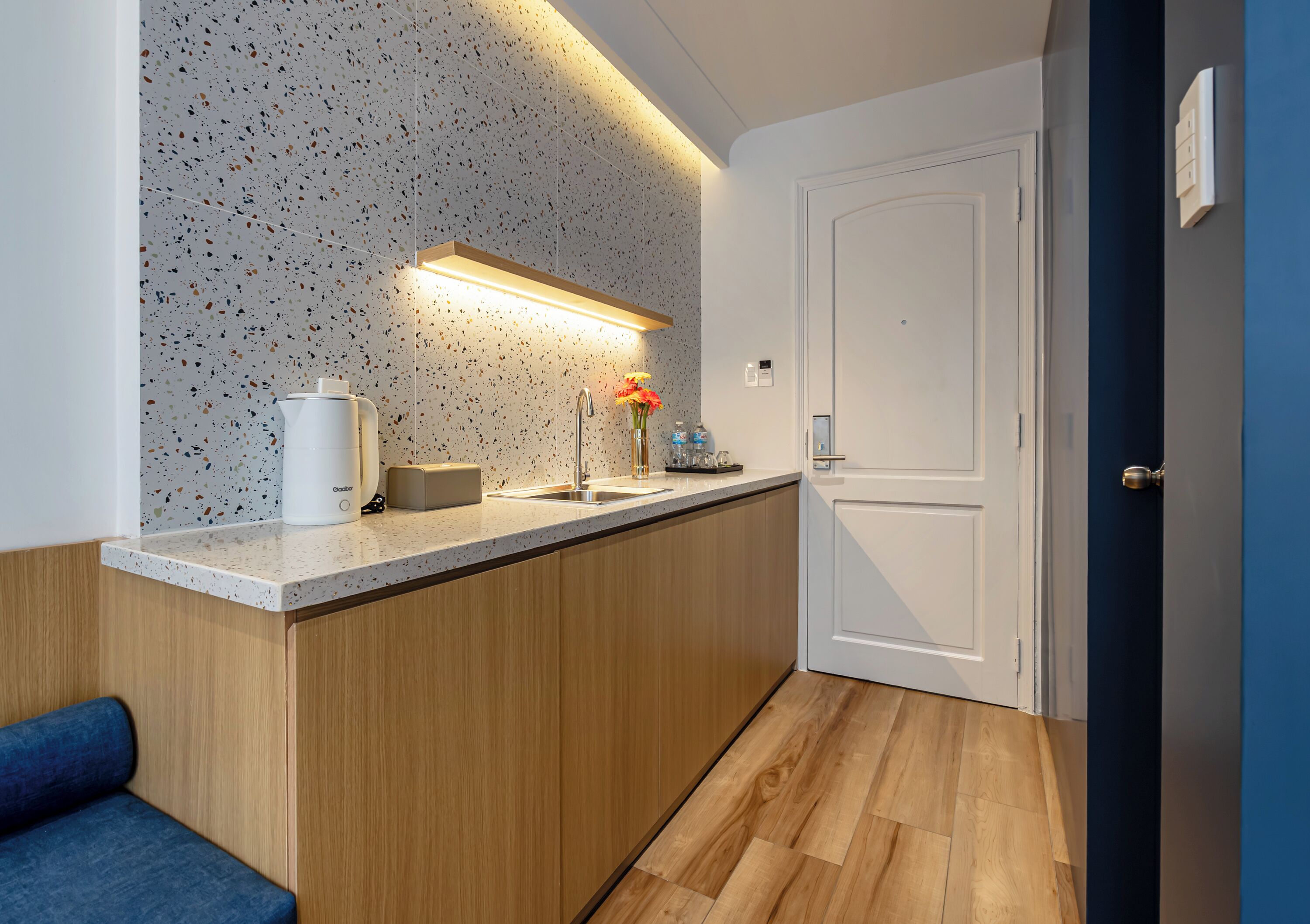 Eine moderne Küche mit geflecktem Fliesenspiegel, Holzschränken, einer Arbeitsplatte, einer Kaffeemaschine, einer Vase mit Blumen und einer Tür rechts.