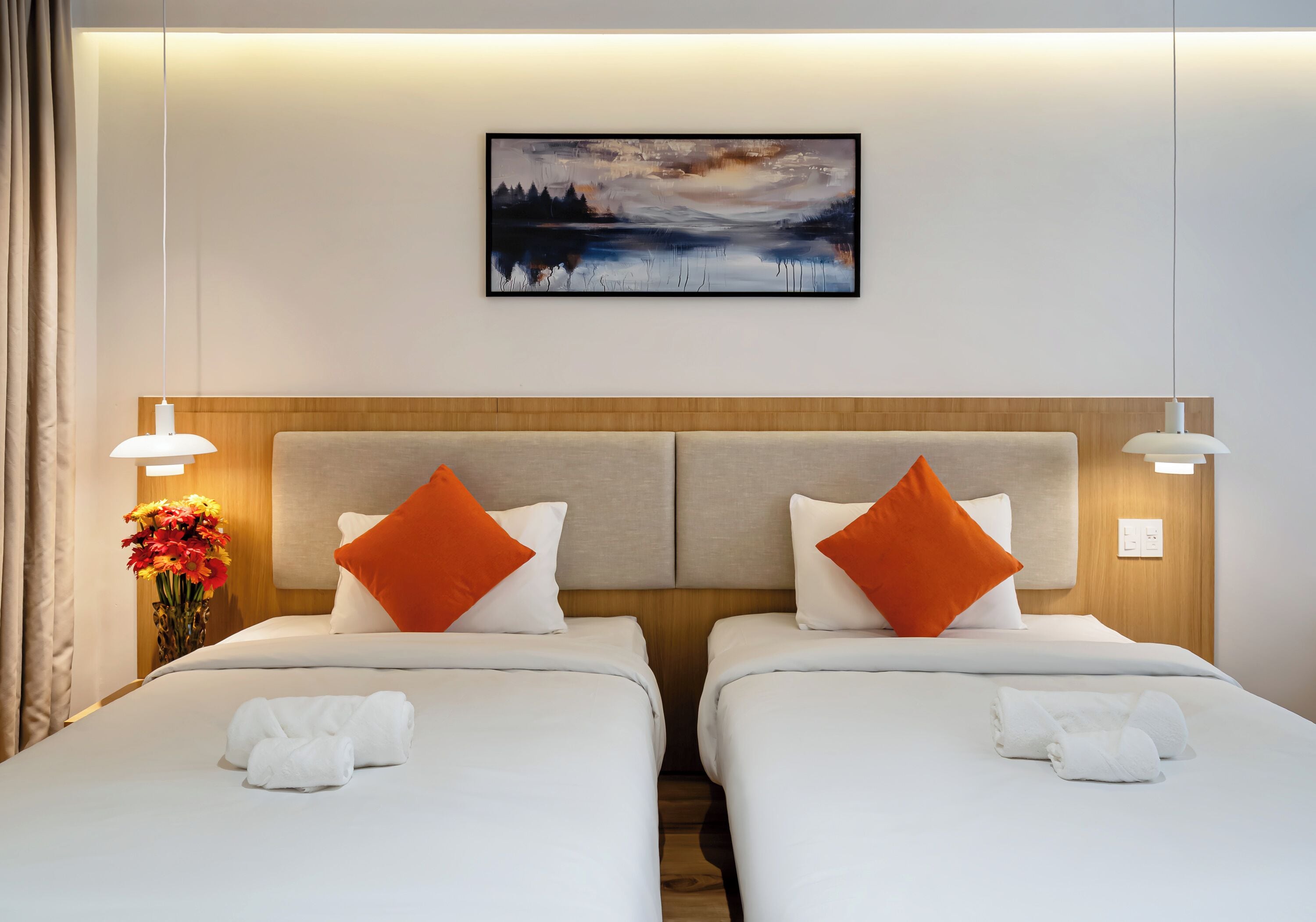 A hotel room with two beds, each with white linens and orange pillows, a wooden headboard, and a framed landscape painting above.
