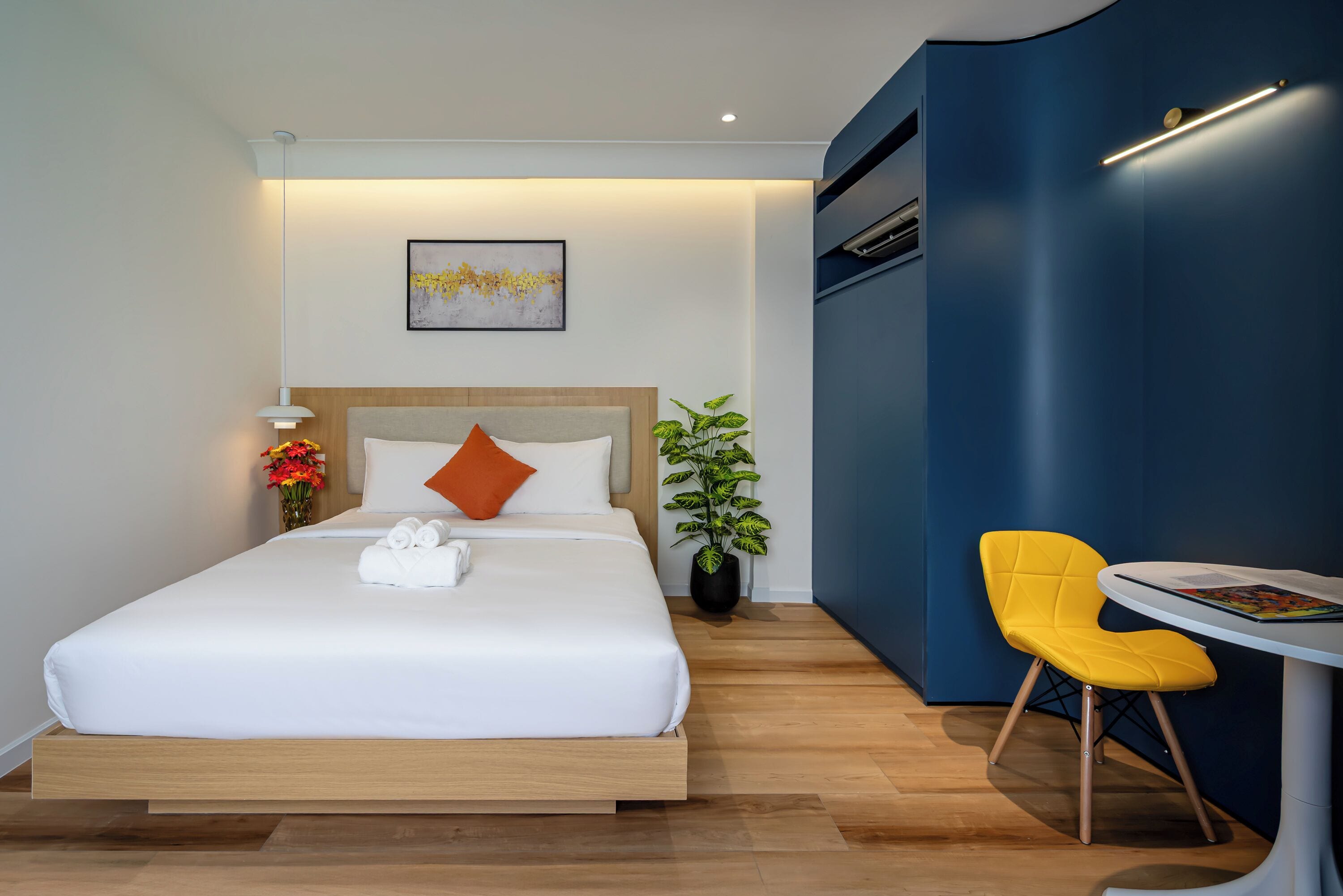 A modern hotel room with a bed, a small table, a chair, and a potted plant.