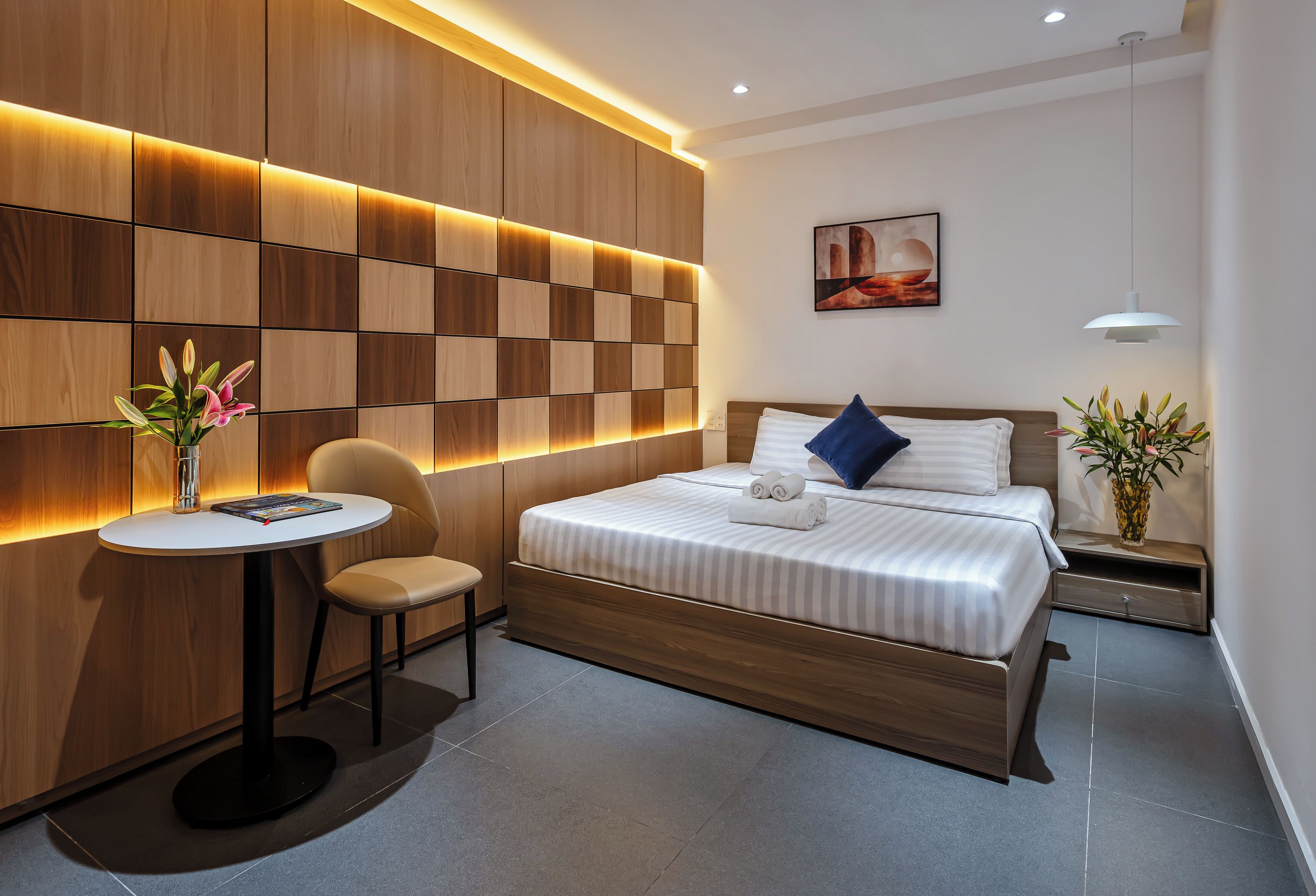 A modern hotel room with a bed, bedside table, chair, and a wall with illuminated panels.