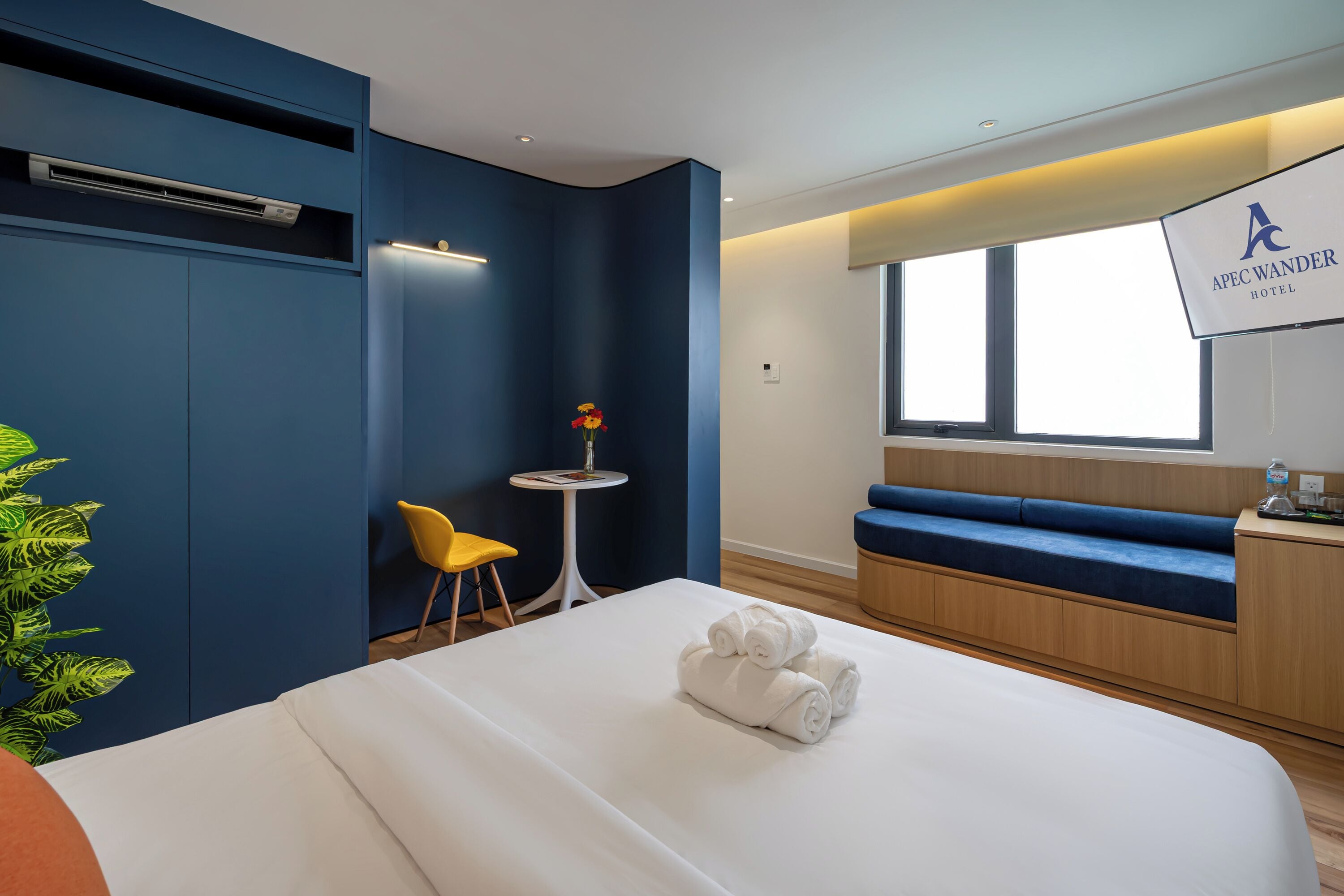 Ein Hotelzimmer mit einem Bett, einem kleinen Tisch mit Blumen, einem gelben Sessel, einem blauen Sofa und einem an der Wand befestigten Fernseher.
