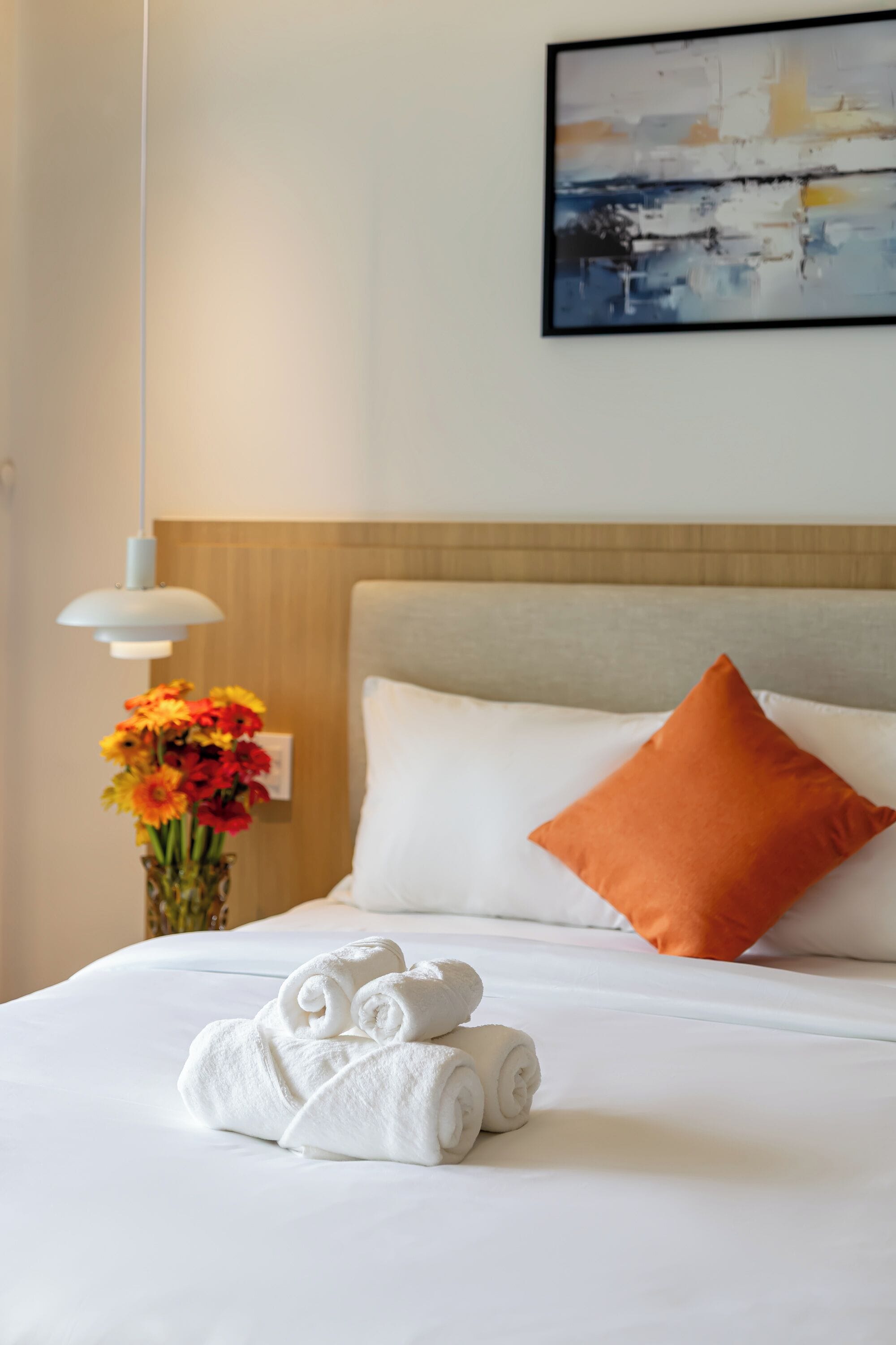 Una cama bien hecha con sábanas blancas, una toalla enrollada y un florero con flores.