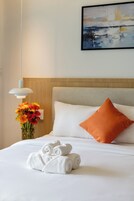Cama bem arrumada com lençóis brancos, uma toalha enrolada e um vaso de flores.