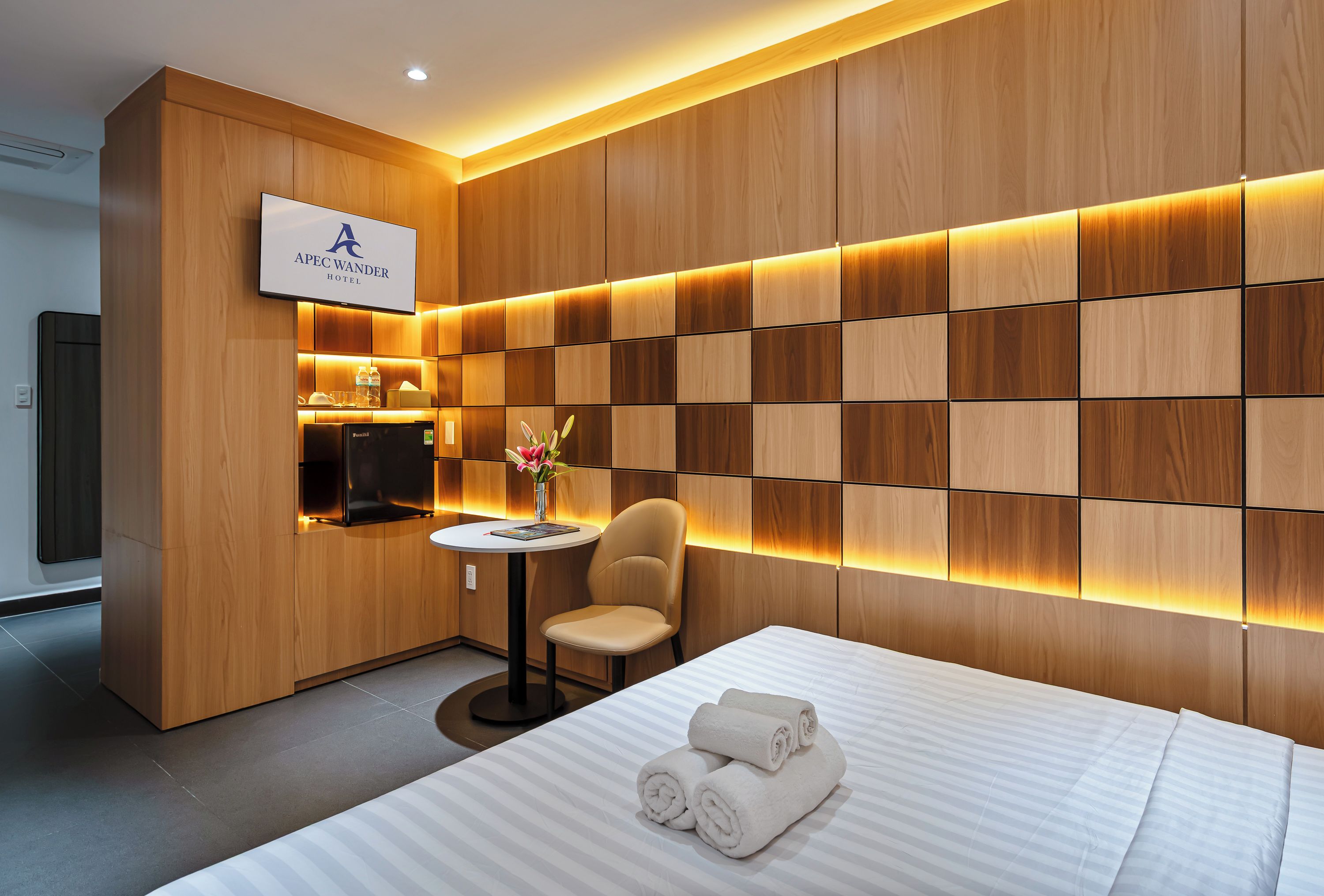 A modern hotel room with a wooden panel wall, a flat-screen TV, a small round table with a chair, and a bed with neatly folded towels.