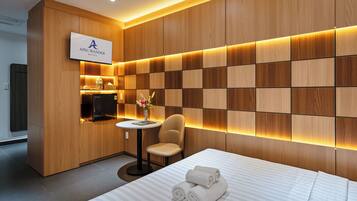 A modern hotel room with a wooden panel wall, a flat-screen TV, a small round table with a chair, and a bed with neatly folded towels.