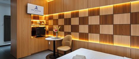 Una habitación de hotel moderna con un panel de pared de madera, un televisor de pantalla plana, una pequeña mesa redonda con una silla y una cama con toallas ordenadamente dobladas.