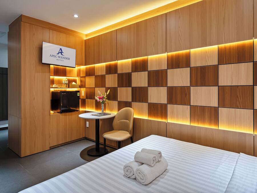 A modern hotel room with a wooden panel wall, a flat-screen TV, a small round table with a chair, and a bed with neatly folded towels.