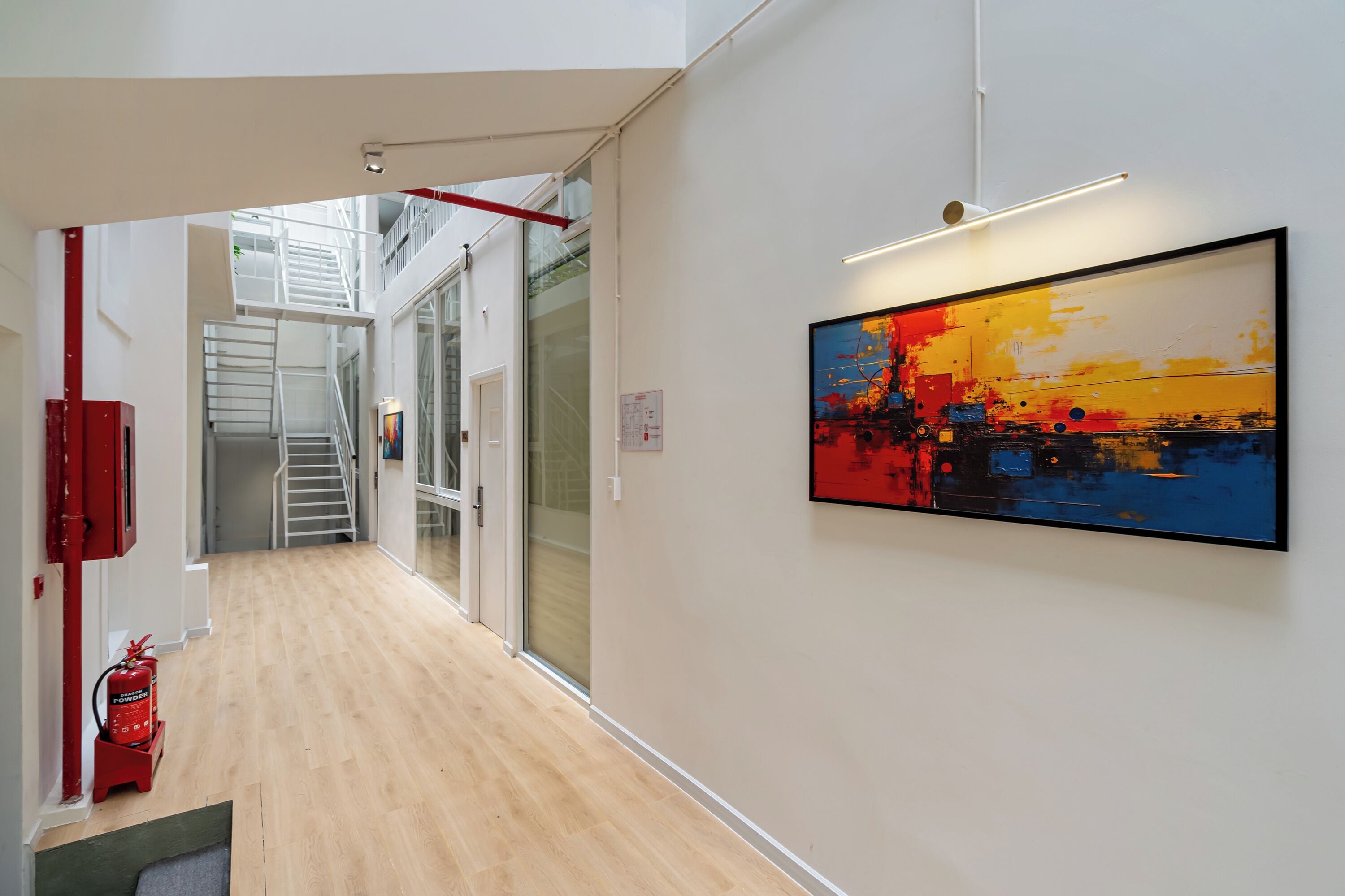 A hallway with a wooden floor, a fire extinguisher, a staircase, and a framed abstract painting.