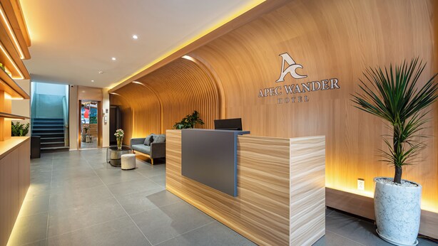 Een moderne hotelontvangst met een houten receptiebalie, een zithoek met een bank en stoelen, en een potplant.