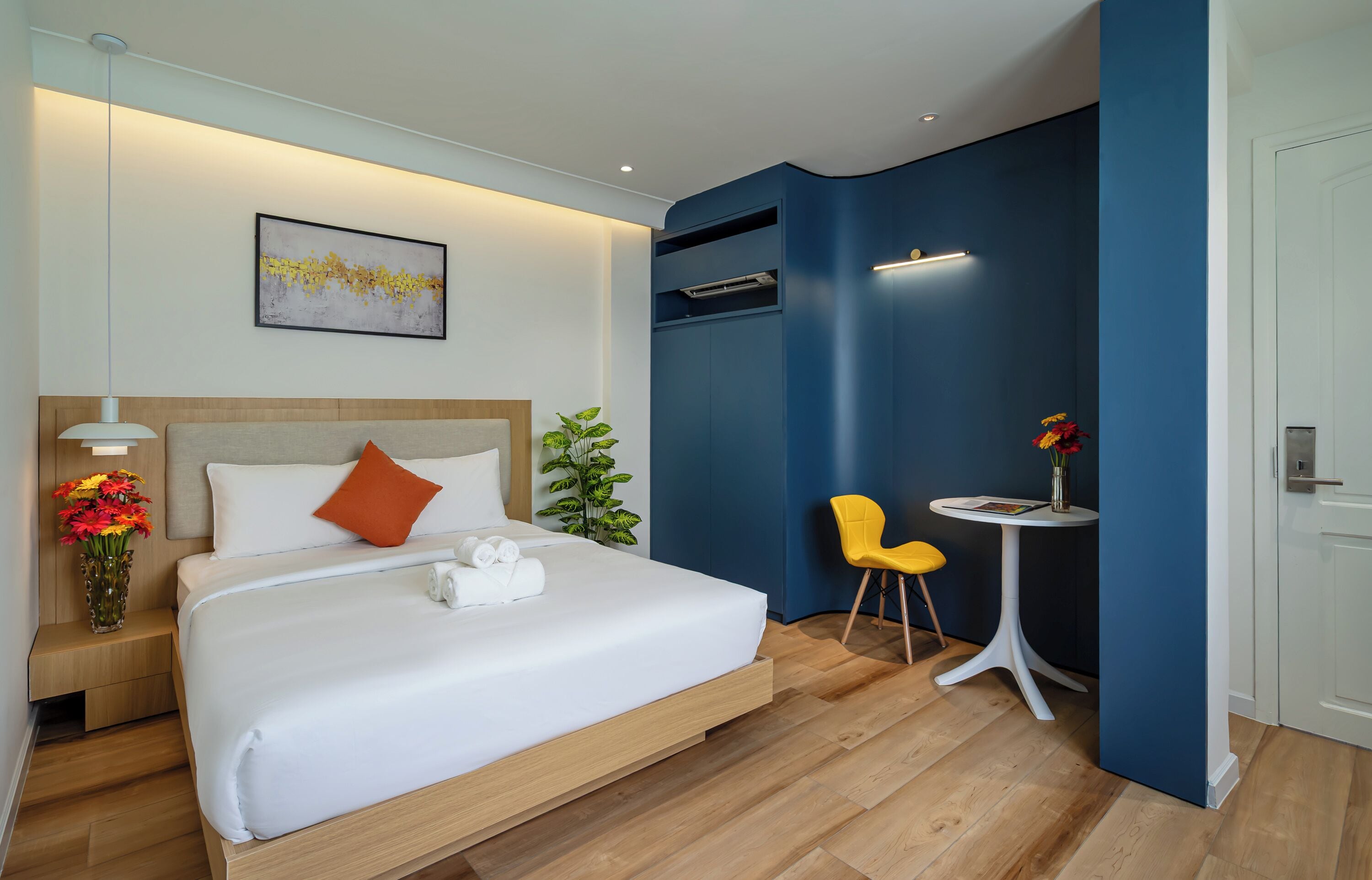 A modern hotel room with a large bed, a bedside table with flowers, a small round table with a chair, and a wall-mounted air conditioning unit.