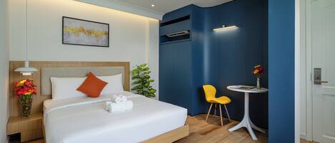 A modern hotel room with a large bed, a bedside table with flowers, a small round table with a chair, and a wall-mounted air conditioning unit.
