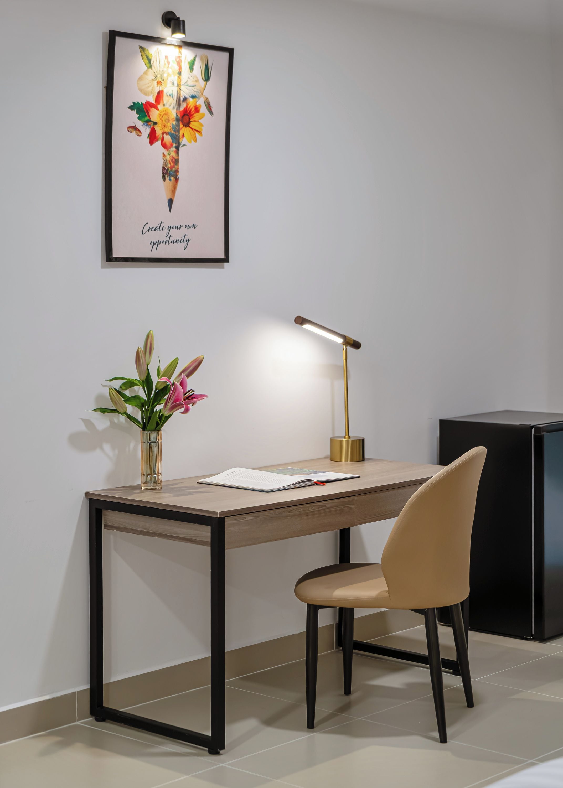 A desk with a lamp, a chair, a vase with flowers, and a framed picture on the wall.