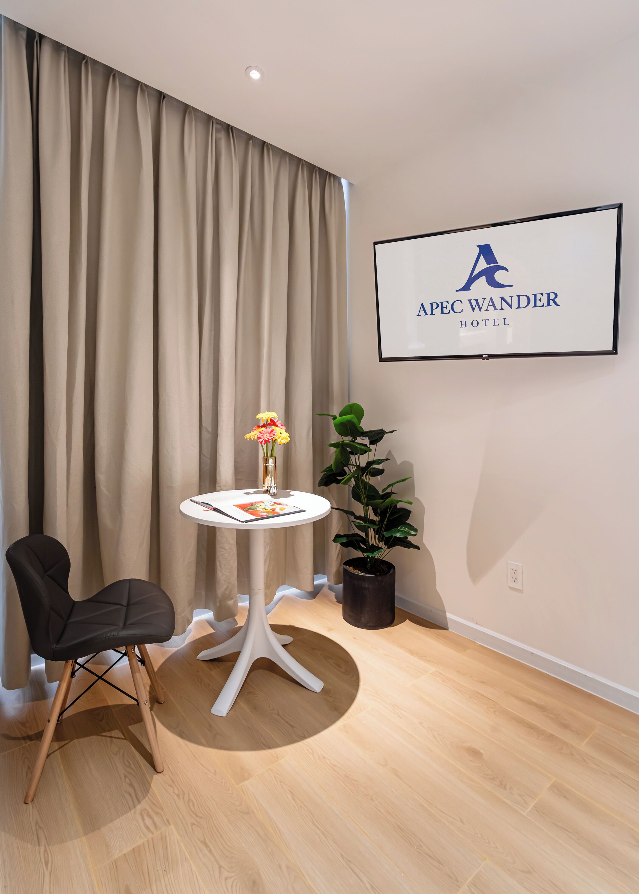 A hotel room with a bed, a chair, a small table with a vase of flowers, a potted plant, and a framed sign on the wall.
