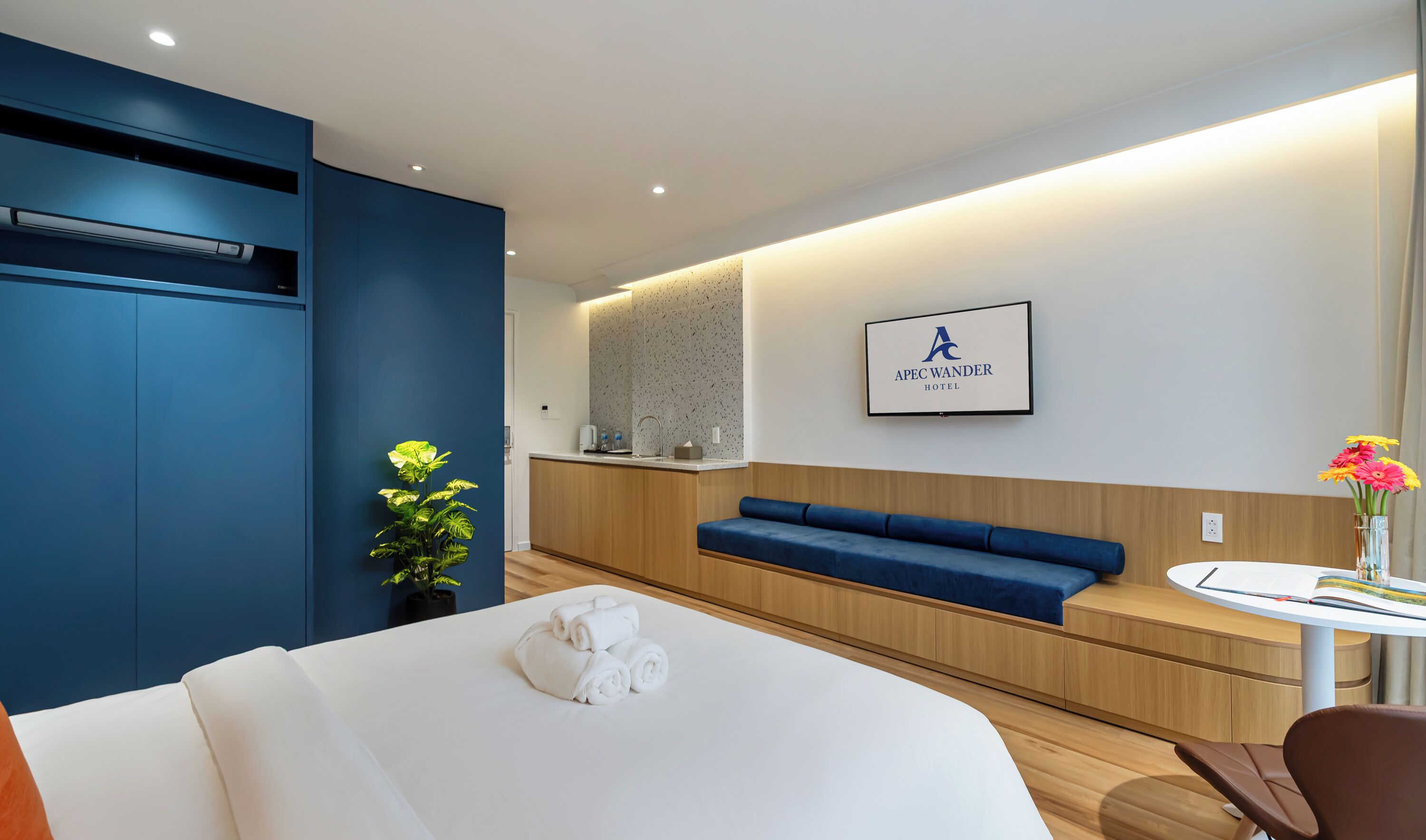 A modern hotel room with a bed, a blue sofa, a small table with a book and flowers, and a kitchenette in the background.