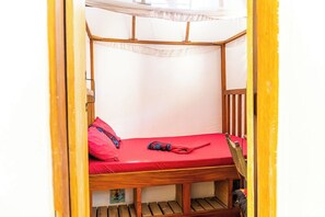 Ein Einzelbett mit roter Matratze und Holzrahmen, einem weißen Baldachin und einer hölzernen Sitzbank am Fußende.