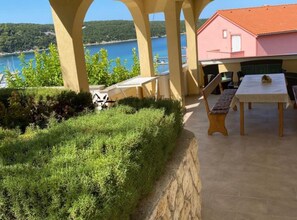 Ein überdachter Außenbereich mit Tisch und Stühlen, Blick auf das Meer und ein Gebäude mit rotem Dach.