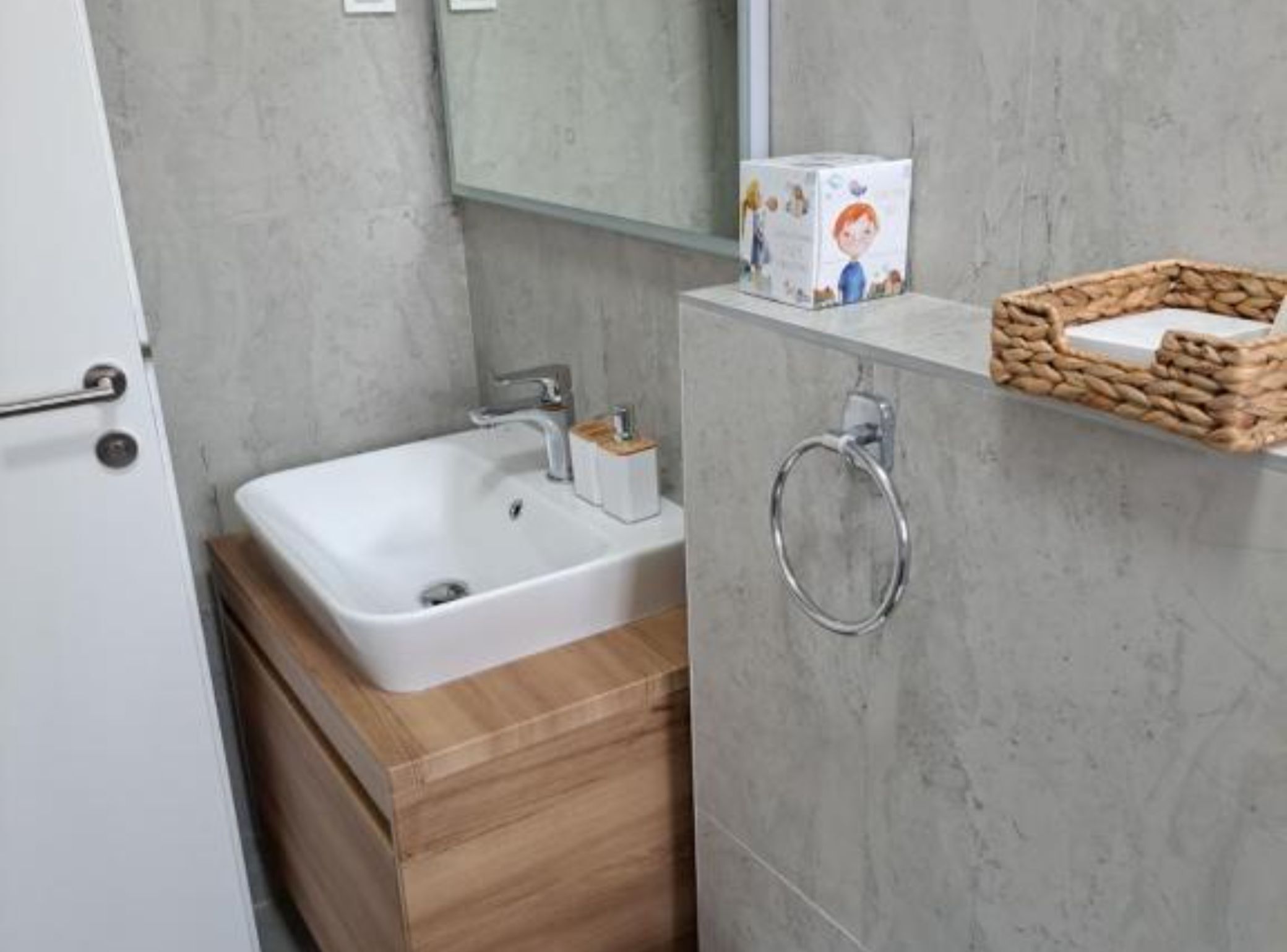 Een badkamer met een witte wastafel, een houten wastafelmeubel, een spiegel en een rieten mand aan de muur.