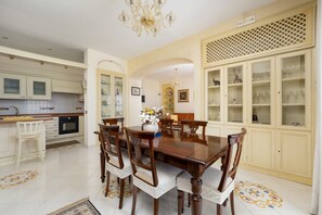 Een eetkamer met een houten tafel en stoelen, een kroonluchter, en een keuken op de achtergrond.