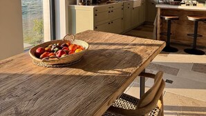 A rustic kitchen with exposed beams, a large window, and a dining area with a woven table and chairs. - Aux Petits Bourgeons (Adainville)
