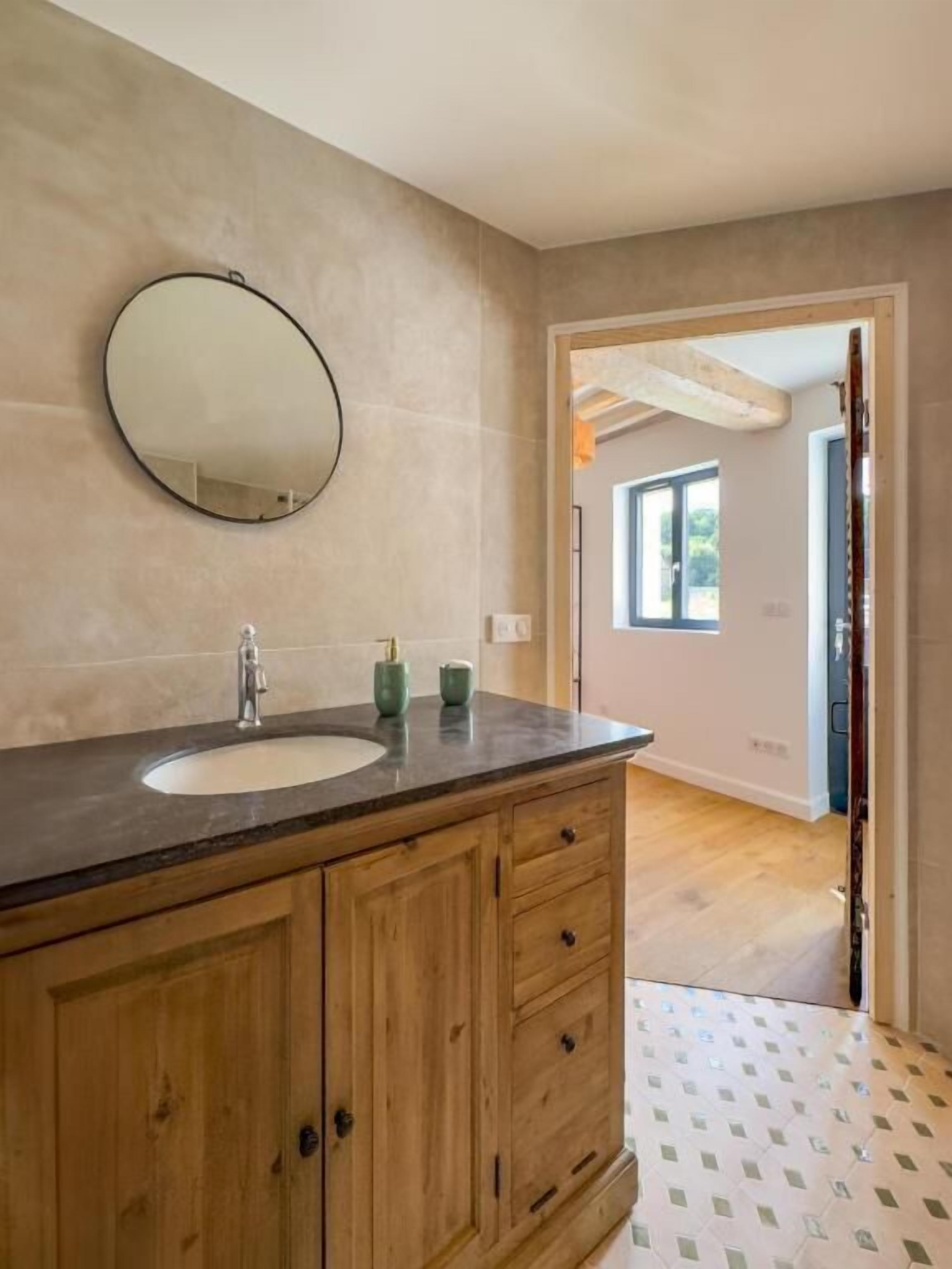 Un baño con tocador de madera, un espejo redondo y una ventana.