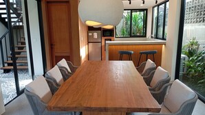 Un espace repas moderne avec une grande table en bois entourée de chaises en osier, une cuisine intégrée avec un réfrigérateur, et une vue sur un jardin grâce à de grandes fenêtres.