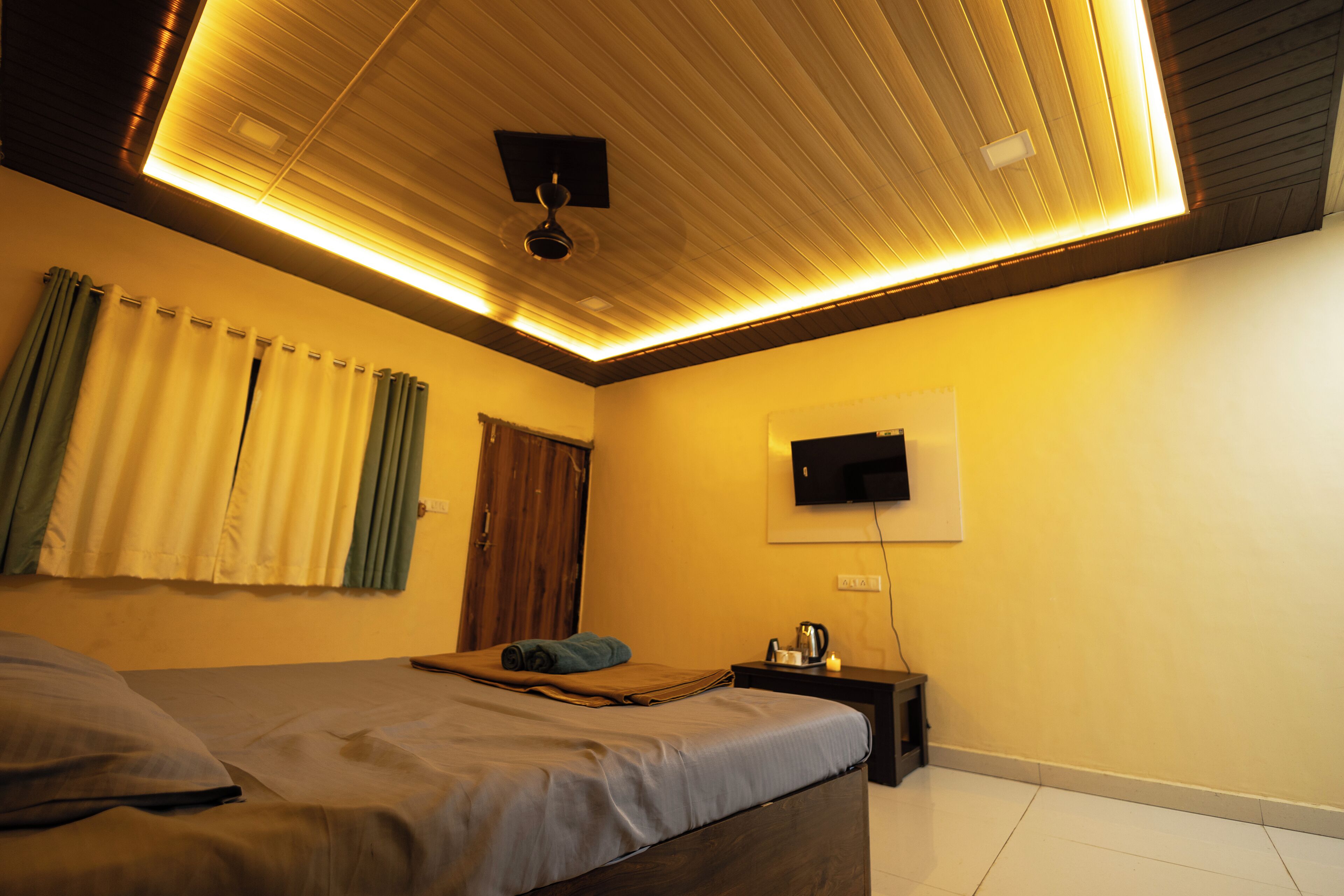 A bedroom with a bed, a ceiling fan, a flat-screen TV mounted on the wall, and a bedside table with a kettle.