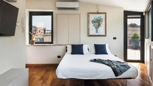 Un dormitorio con una cama grande, una ventana con vistas a edificios, un equipo de aire acondicionado y un cuadro en la pared.