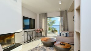 Un salon moderne dotĂ© dâun tĂ©lĂ©viseur Ă Ă©cran plat, dâune cheminĂ©e, dâun canapĂ© et dâun balcon offrant une vue sur la mer.