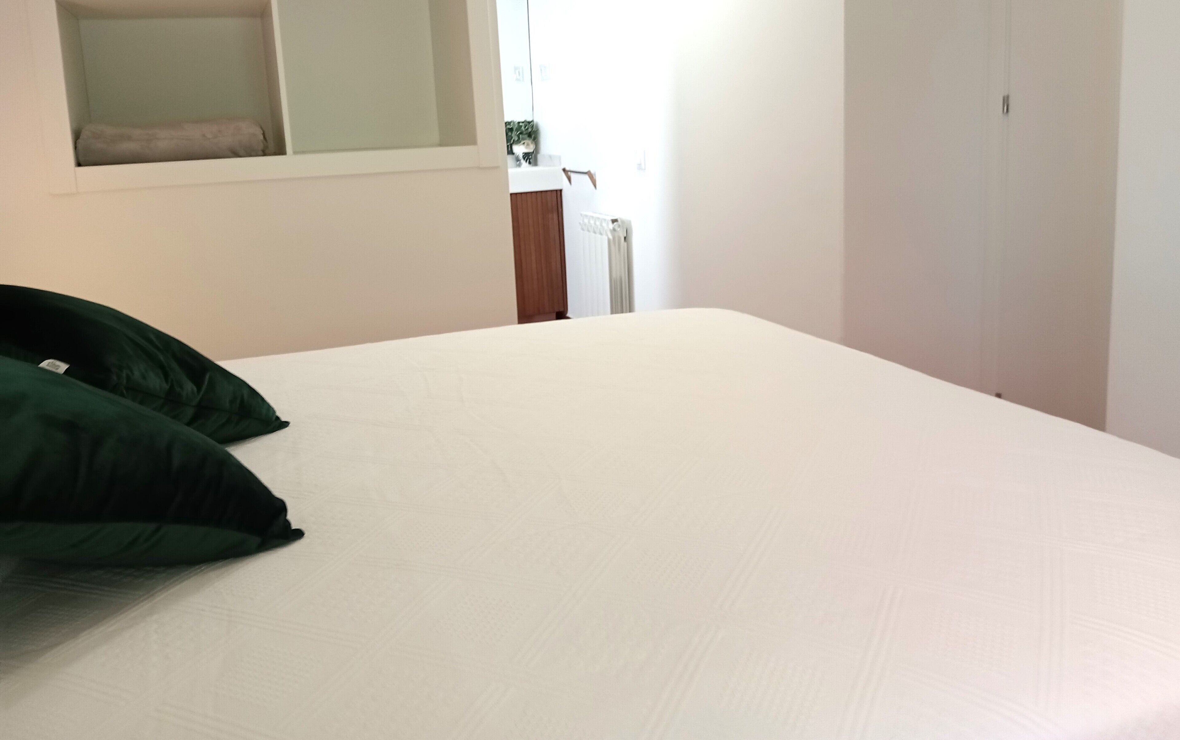 Una cama bien tendida con sábanas blancas y dos almohadas verdes, una mesita de noche de madera con una planta, y un radiador blanco al fondo.