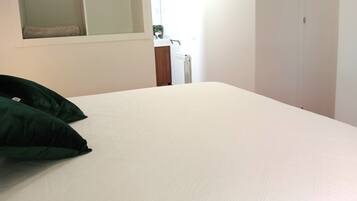 Una cama bien tendida con sábanas blancas y dos almohadas verdes, una mesita de noche de madera con una planta, y un radiador blanco al fondo.