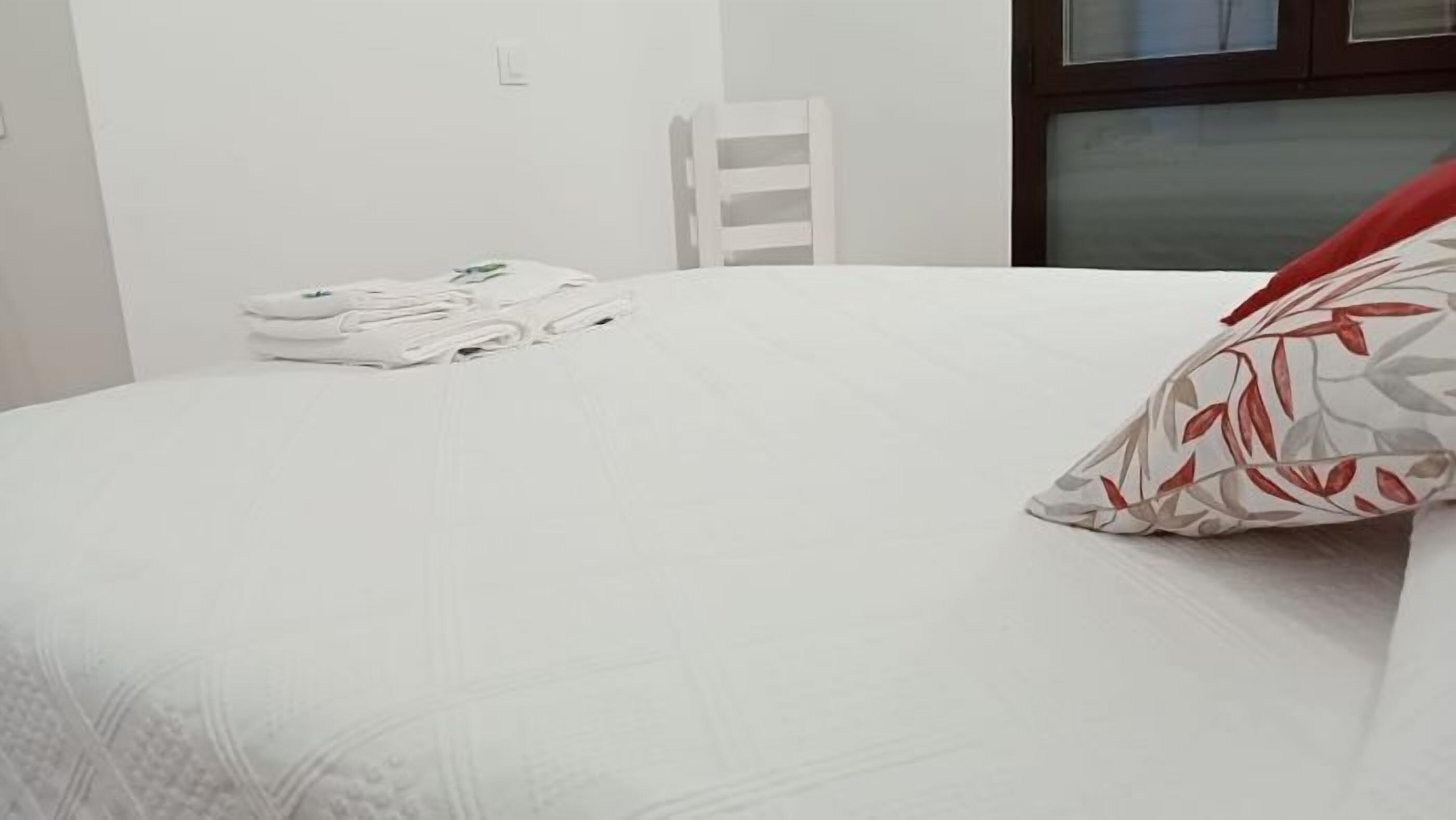 A neatly made bed with white bedding and a red and grey patterned pillow. There is a white chair and a stack of white towels in the background.