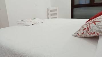 A neatly made bed with white bedding and a red and grey patterned pillow. There is a white chair and a stack of white towels in the background.