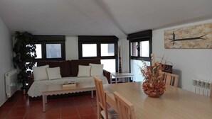 Una sala de estar acogedora con un sofá, una mesa de comedor y sillas. Hay un ventanal con persianas, un radiador y un cuadro en la pared.