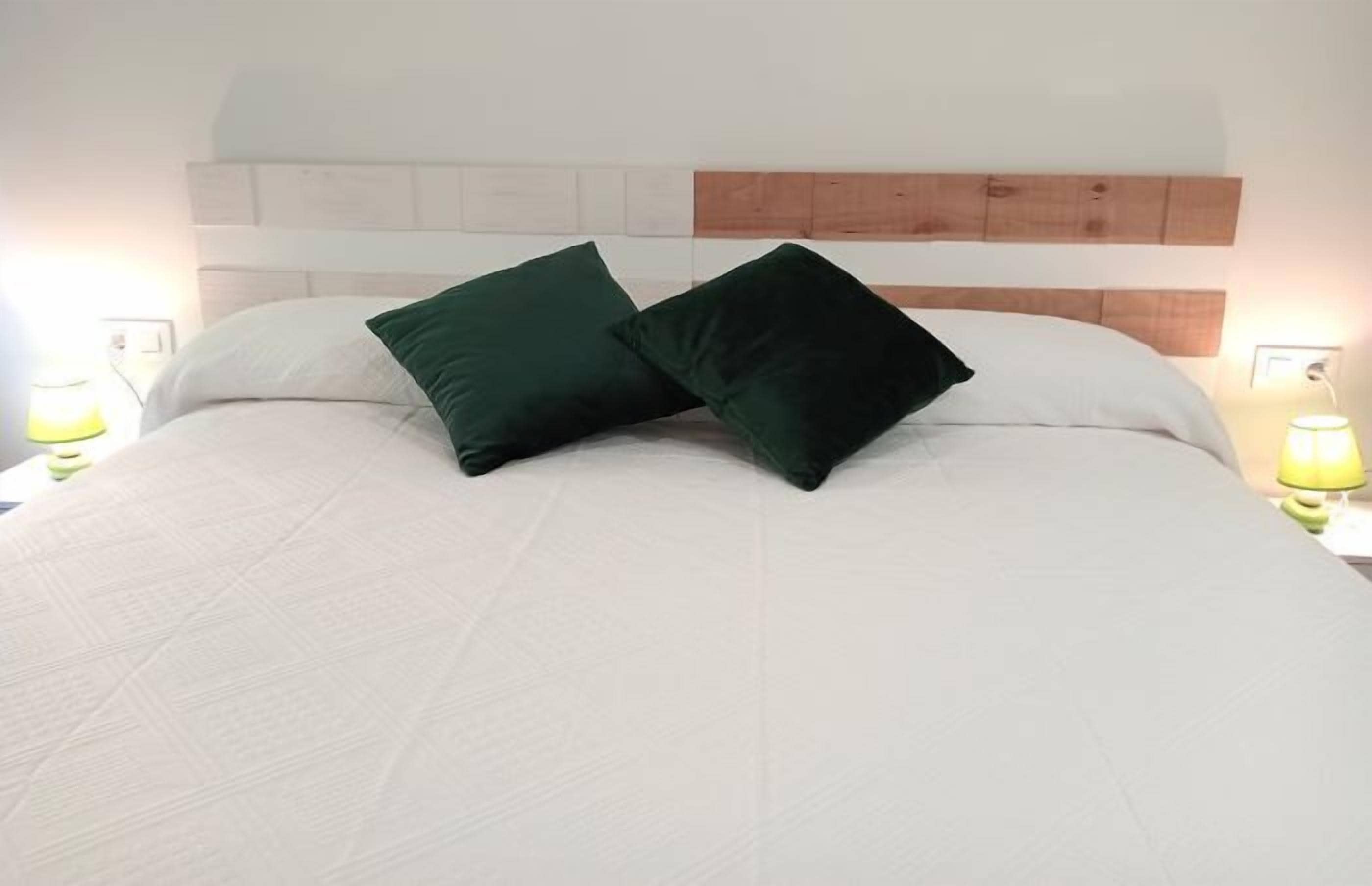 A bed with white bedding and two dark green pillows, a wooden headboard, and two bedside lamps with yellow shades.
