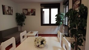 Un comedor con una mesa blanca, sillas blancas, un jarrón blanco con flores, un sofá gris y dos plantas en macetas.