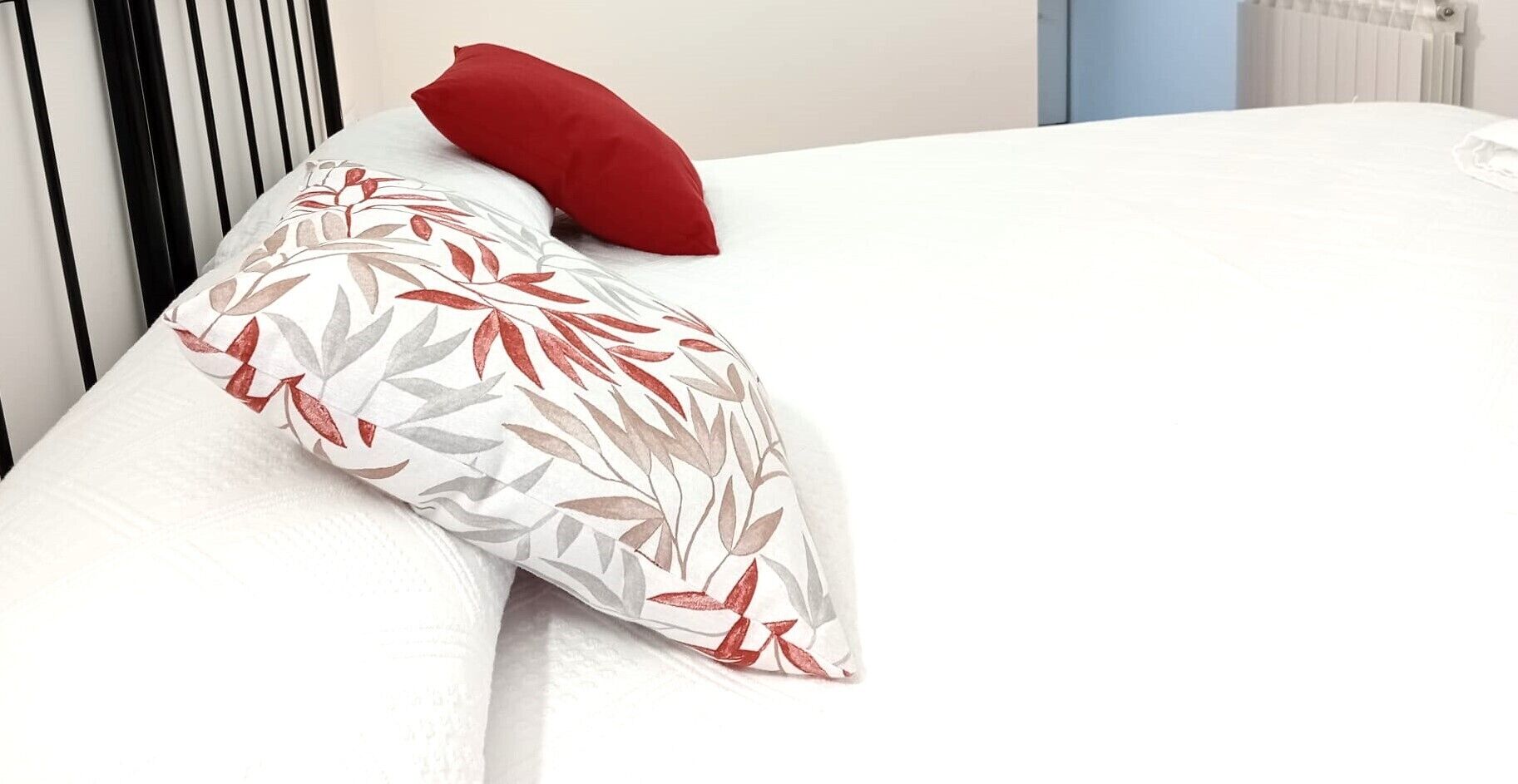 Una cama con un almohadón blanco con estampado de hojas y un almohadón rojo.
