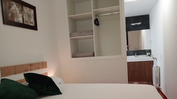 Un dormitorio con cama, almohadas, armario, espejo y baño con lavabo y toallero.