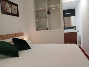 Een slaapkamer met een bed, kussens, een kledingkast, een spiegel en een badkamer met een wastafel en handdoekhouder.