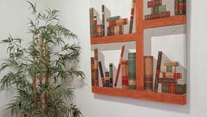 Una habitación con una amplia planta de bambú en maceta y un cuadro colgado en la pared que muestra libros y formas geométricas.