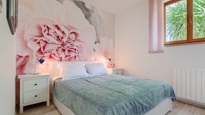 Quarto com um grande mural floral, cama com manta azul de tricô, duas luminárias de cabeceira e janela com vista para vegetação.