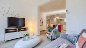 A modern living room with a flat-screen TV, a white entertainment unit, a colorful sofa, and a dining area in the background.