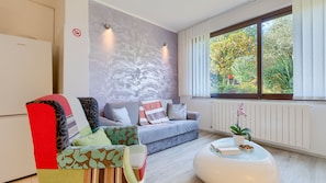 A modern living room with a sofa, armchair, and ottoman, a large window with a view of greenery, and a wall-mounted radiator.