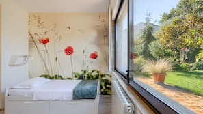 A bedroom with a bed, a wall-mounted light, and a window overlooking a garden.