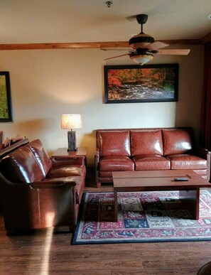 A living room with a brown leather sofa, a matching armchair, a wooden coffee table, a floor lamp, and a ceiling fan.