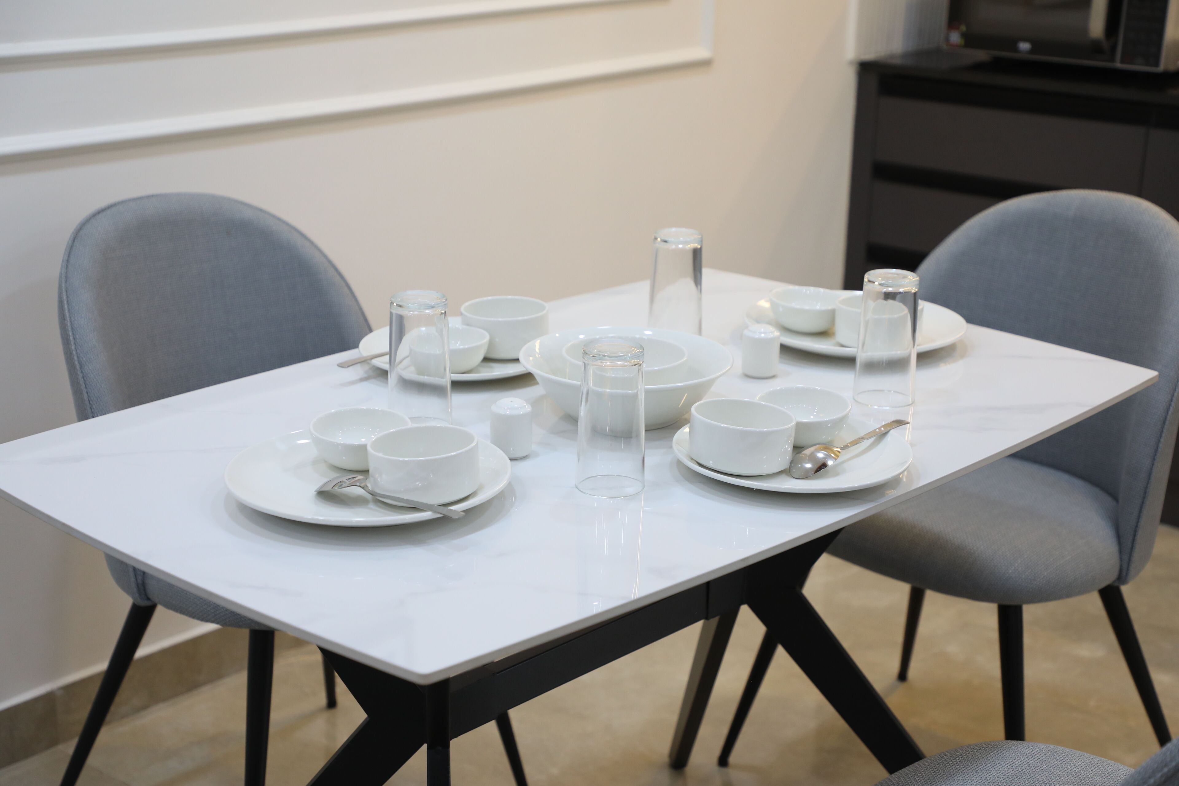 A dining table set with white plates, bowls, and cutlery, surrounded by grey chairs.