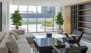 A modern living room with a sofa, coffee table, and chairs, large windows, and indoor plants.