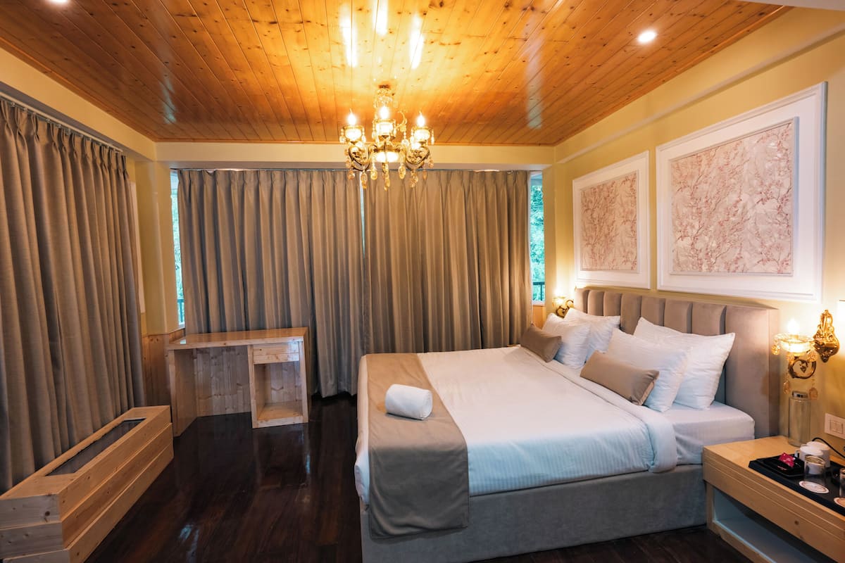 A hotel room with a wooden ceiling, a large bed with a headboard, a nightstand with a lamp, and a window with curtains.