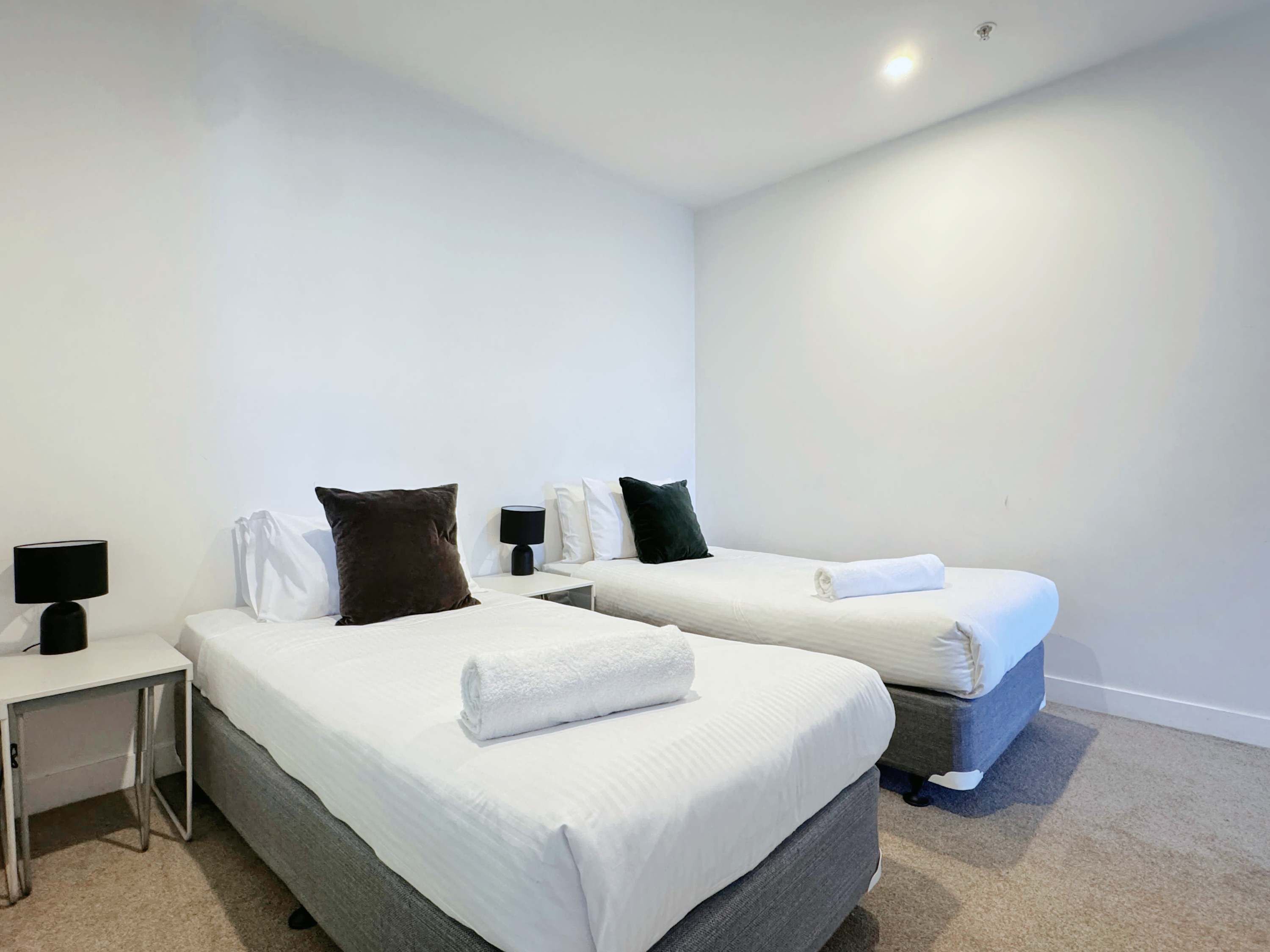 Habitación de hotel con dos camas, cada una con ropa de cama y almohadas blancas, una mesita de noche pequeña con lámpara y suelo alfombrado.